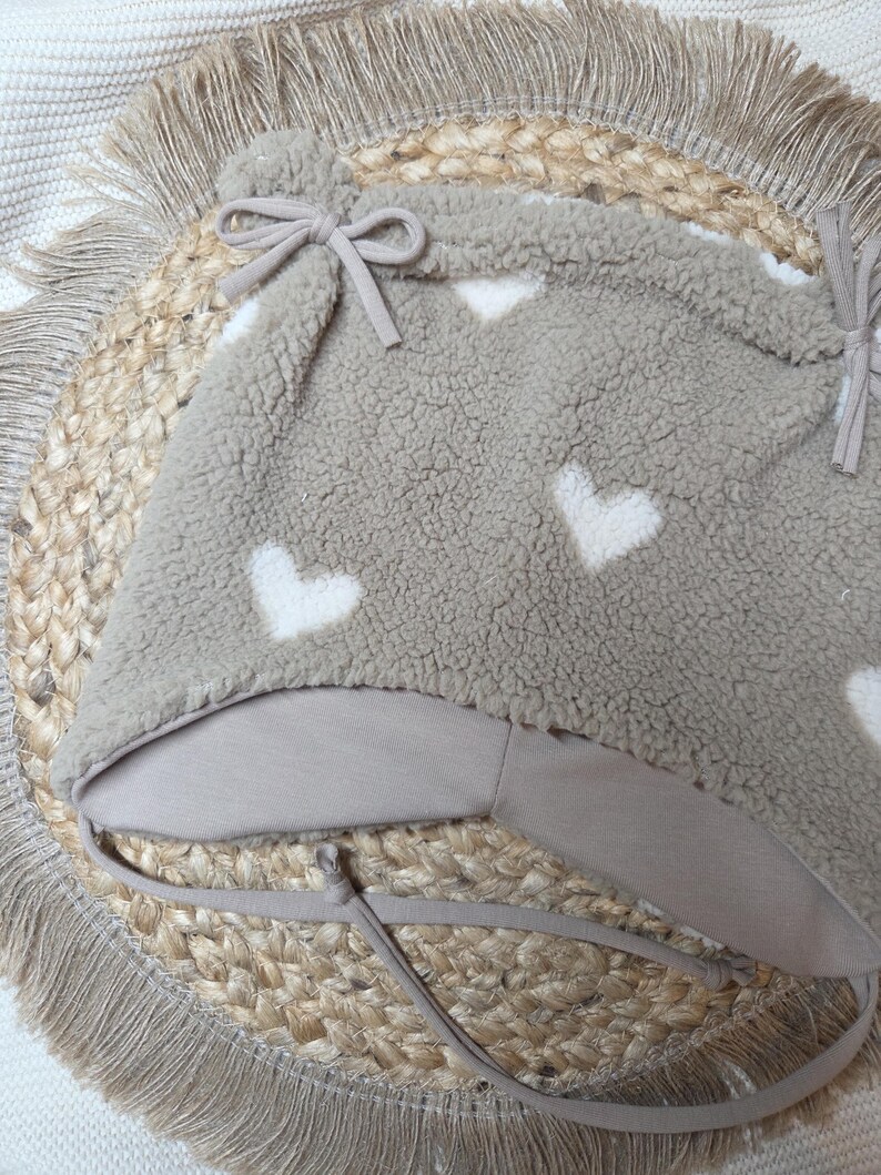May include: A taupe baby hat with white heart patterns. The hat is made of a soft, textured material and has a chin strap with a bow. It rests on a round, woven mat with a fringed edge. A cosy accessory.
