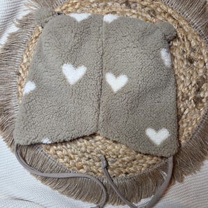 May include: A taupe fleece hat featuring white heart patterns. The hat has ear flaps and ties for fastening. Designed for warmth, it's suitable for cold weather. The hat is displayed on a woven, circular mat. The hat is made of a soft, fluffy material.
