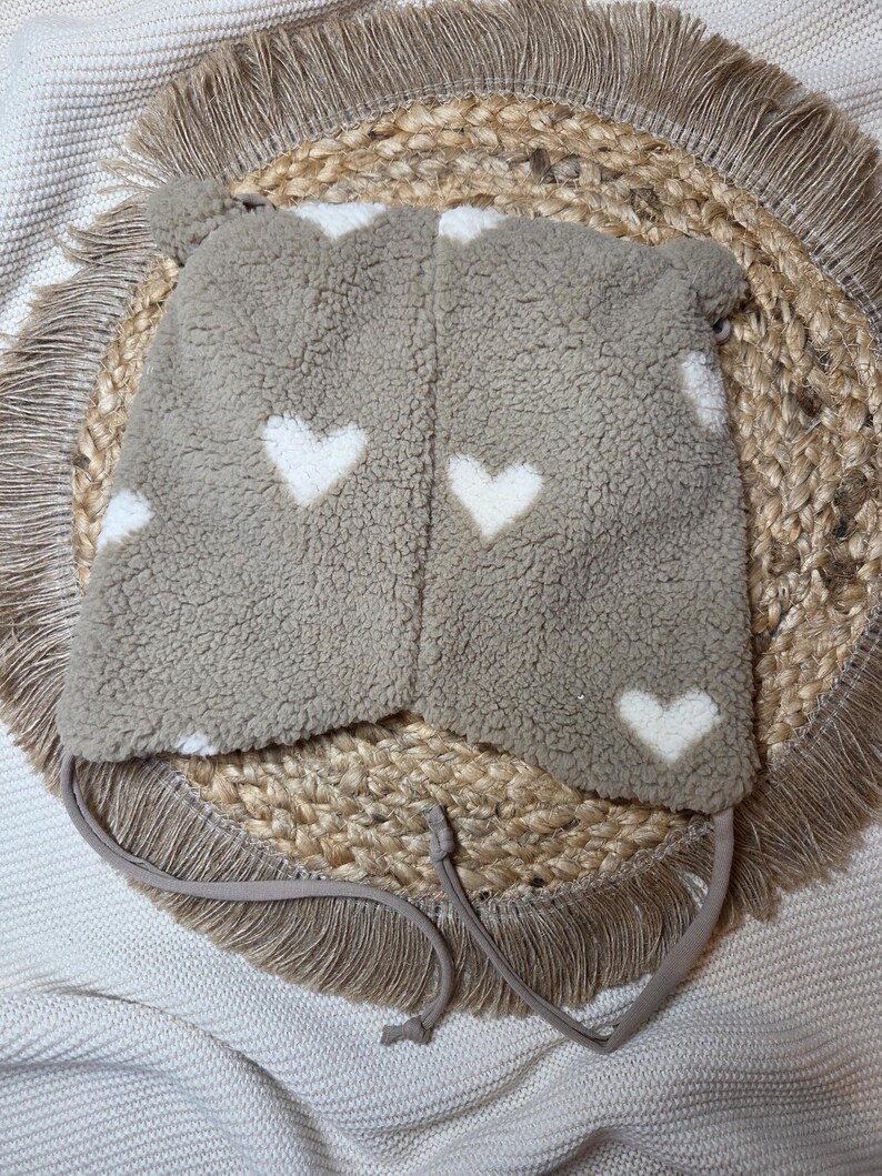 May include: A taupe fleece hat featuring white heart patterns. The hat has ear flaps and ties for fastening. Designed for warmth, it's suitable for cold weather. The hat is displayed on a woven, circular mat. The hat is made of a soft, fluffy material.