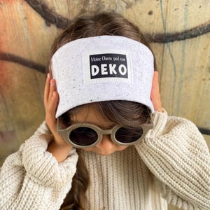 Puede incluir: Una visera blanca con motas negras y el texto "Meine Ohren sind nur DEKO" en un rectángulo negro. La visera se usa con gafas de sol y un suéter color crema.