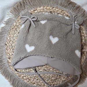May include: A taupe baby hat with white heart patterns. The hat is made of a soft, textured material and features ear flaps and ties. The hat is placed on a woven, circular mat with a fringed edge.