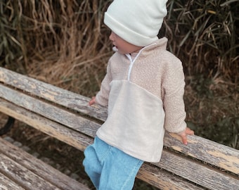 May include: A young child wearing a white beanie, a beige fleece pullover with a zipper, and blue jeans sits on a wooden bench.