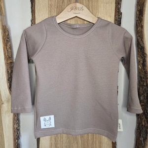 Könnte beinhalten: Ein langärmeliges, taupefarbenes Shirt mit gerippter Struktur, das an einem Holzbügel hängt. Ein kleines weißes quadratisches Patch mit einer Hasenillustration ist auf der unteren linken Seite aufgenäht. Der Bügel trägt den Text "JmLa HANDMADE".