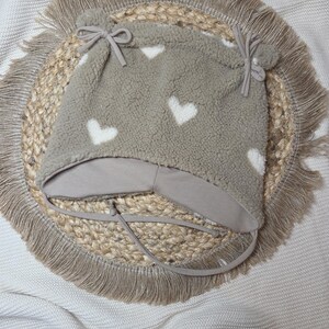 May include: A grey fleece hat featuring white heart shapes. The hat has ear flaps and ties, and is decorated with two small pom-poms. The hat is designed for babies and young children.