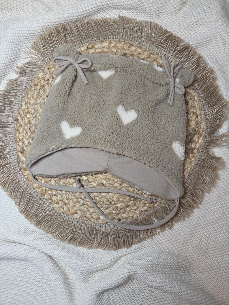 May include: A grey fleece hat featuring white heart shapes. The hat has ear flaps and ties, and is decorated with two small pom-poms. The hat is designed for babies and young children.