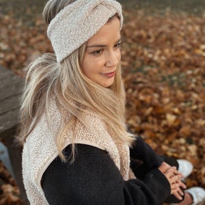 Könnte beinhalten: Eine Frau trägt ein beiges Fleece-Stirnband und eine beige Fleece-Weste über einem schwarzen Langarmshirt. Sie sitzt auf einer Holzbank mit Herbstlaub im Hintergrund.