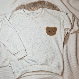 Könnte beinhalten: Ein weißer Pullover mit einem braunen Bären-Applikationen auf der Vorderseite. Der Pullover hat ein gesprenkeltes Muster.
