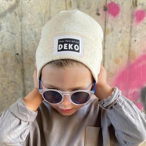 Peut inclure: Un bonnet beige à motifs mouchetés avec un écusson rectangulaire portant l'inscription "Meine Elven sind nur DEKO" en noir. Le bonnet est porté par une personne portant des lunettes de soleil à monture transparente et branches bleues.