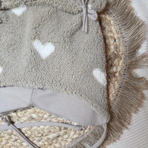 May include: A taupe baby hat with white heart patterns. The hat is made of a soft, textured material and has a light brown lining and ties. The hat is placed on a woven, circular mat with a fringed edge.