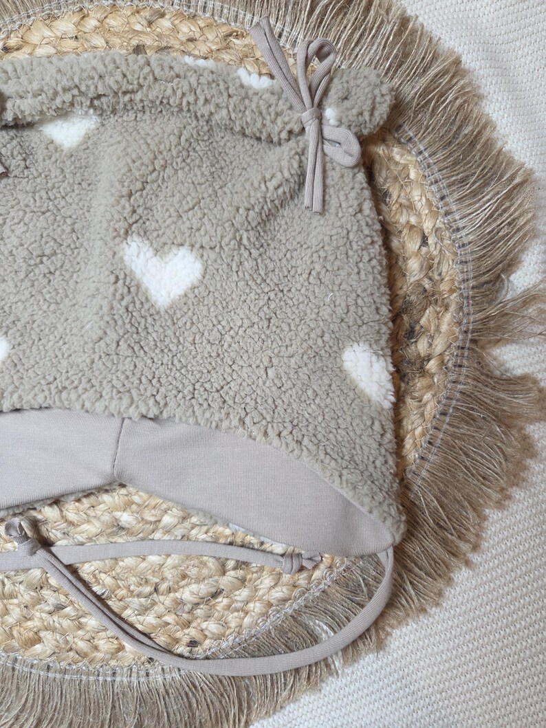 May include: A taupe baby hat with white heart patterns. The hat is made of a soft, textured material and has a light brown lining and ties. The hat is placed on a woven, circular mat with a fringed edge.