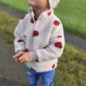 Peut inclure: Une veste en polaire crème et duveteuse ornée de taches rouges ressemblant à des coccinelles. La veste a une fermeture à demi-zip et des poignets côtelés beiges. L'enfant porte un jean bleu et un bonnet gris clair.