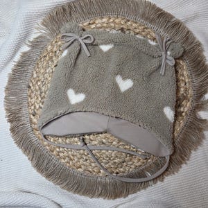 May include: A grey fleece baby hat patterned with white hearts. The hat features a light grey brim and ties, and is decorated with small bows and pom-poms. It is displayed on a woven jute placemat with a fringed edge.