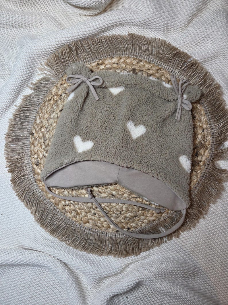 May include: A grey fleece baby hat patterned with white hearts. The hat features a light grey brim and ties, and is decorated with small bows and pom-poms. It is displayed on a woven jute placemat with a fringed edge.