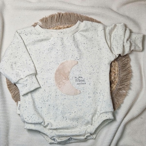 Könnte beinhalten: Ein weißer Baby-Strampler mit einem gesprenkelten Muster und einer beigefarbenen Filz-Mond-Applikation. Der Strampler hat den Text "to the moon and back" aufgedruckt.