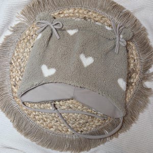 May include: A taupe baby hat with white heart patterns. The hat has ear flaps, ties, and a brim, made from a soft, textured material. It rests on a round placemat with a fringed edge.