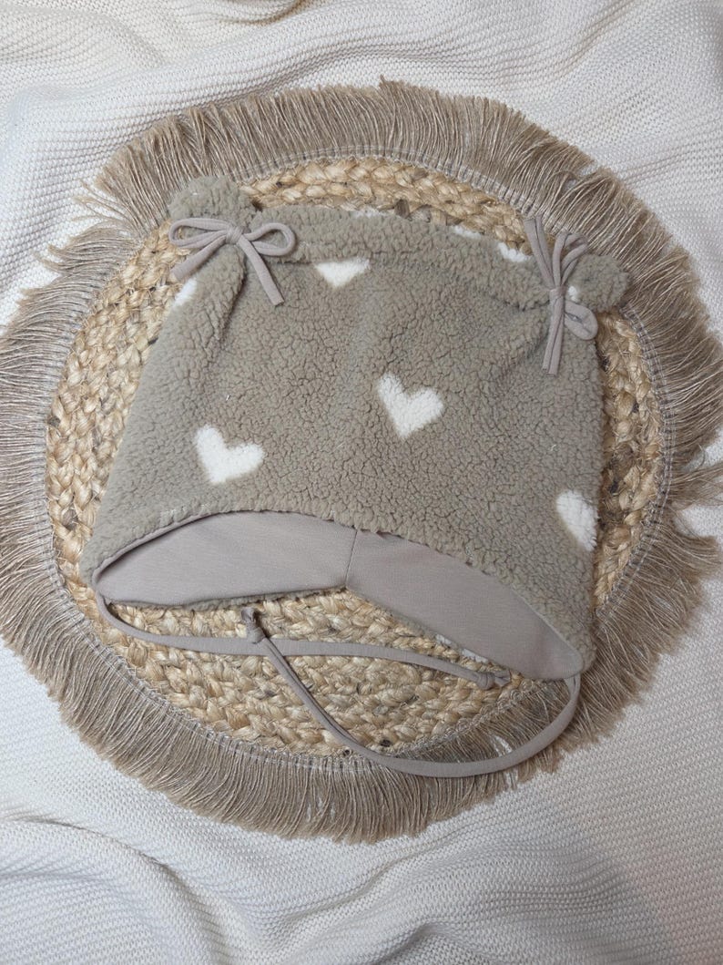 May include: A taupe baby hat with white heart patterns. The hat has ear flaps, ties, and a brim, made from a soft, textured material. It rests on a round placemat with a fringed edge.