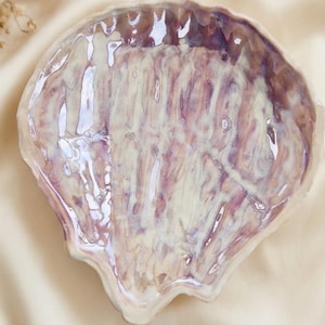 Könnte beinhalten: Eine Keramikschale in Form einer Muschel mit einer glänzenden, irisierenden Glasur. Die Oberfläche weist wirbelnde Muster in Creme, zartem Lavendel und tiefem Violett auf, was ein reflektierendes, marmoriertes Aussehen mit gewelltem Rand erzeugt.