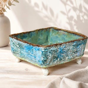 Op de afbeelding: Een vierkante keramische kom met een getextureerd, hobbelig buitenoppervlak met een mix van blauw, turquoise en groen glazuur. De rand heeft een donkerbruine afwerking en de kom rust op vier kleine, witte, afgeronde voetjes op de hoeken.