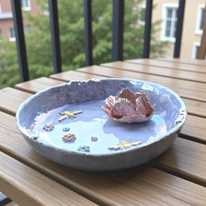 Op de afbeelding: Een keramische kom met een lichtblauwe iriserende glazuur. De kom heeft een roze lotusbloem in het midden en kleine versieringen van bijen en bloemen. De kom staat op een houten tafel.