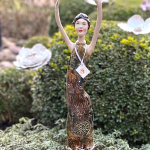 Può includere: Un paletto da giardino in ceramica raffigurante una figura snella con le braccia alzate. La figura indossa un abito fantasia scuro e un berretto scuro. Il paletto è montato su un'asta di metallo e ha un'etichetta al collo.