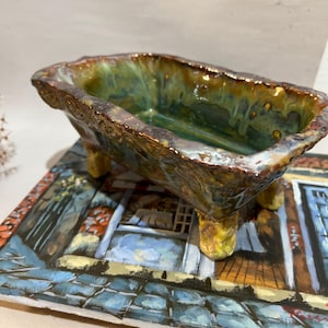 Könnte beinhalten: Eine kleine, rechteckige Keramikschale in Form einer Badewanne mit vier kurzen Beinen. Die Außen- und Innenseite weisen eine glänzende, gesprenkelte Glasur in tiefen Grün-, Braun- und Goldgelbtönen auf, mit einem strukturierten, unebenen Rand.