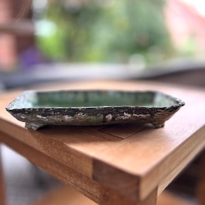Puede incluir: Un plato rectangular de cerámica con interior verde y borde oscuro texturizado. El plato está elevado sobre pequeños pies y descansa sobre una mesa de madera. Los bordes tienen un aspecto envejecido y rústico.
