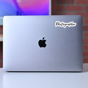 May include: A silver laptop computer with a black Apple logo and a white sticker that says "Photographer" on the top right corner.