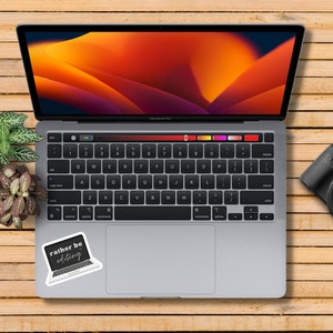 May include: A silver laptop computer with a black keyboard and a sticker that says "rather be editing". The laptop is open and the screen is displaying a colorful abstract design. A black camera is on the wooden surface to the right of the laptop.