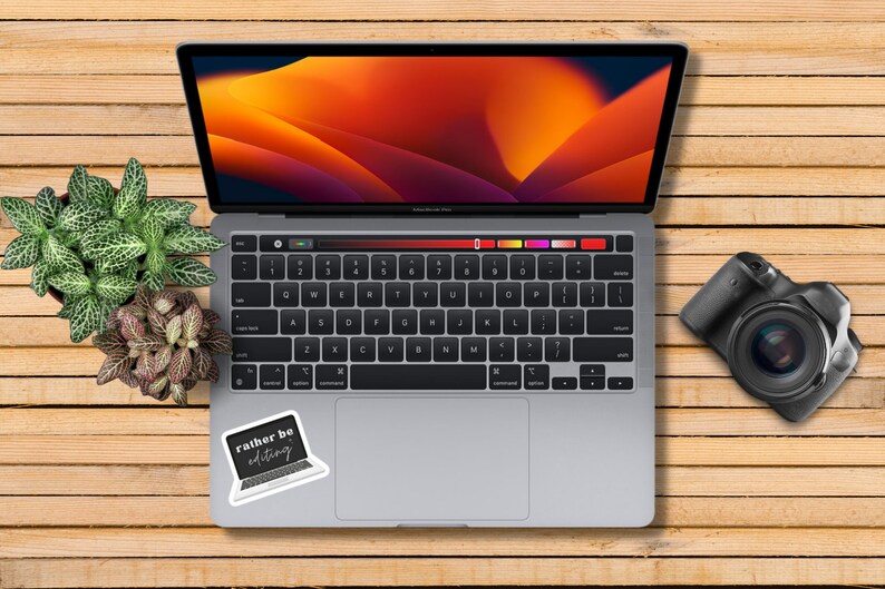 May include: A silver laptop computer with a black keyboard and a sticker that says "rather be editing". The laptop is open and the screen is displaying a colorful abstract design. A black camera is on the wooden surface to the right of the laptop.