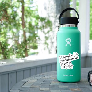 May include: A turquoise Hydro Flask water bottle with a white sticker that reads "Photographer Life 5% Taking Photos 5% Coffee 15% Editing". A black camera is sitting on a gray table next to the bottle.
