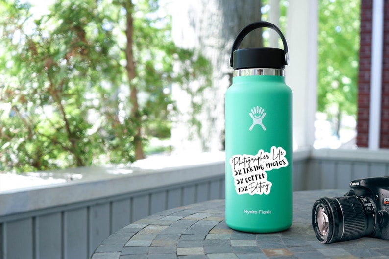 May include: A turquoise Hydro Flask water bottle with a white sticker that reads "Photographer Life 5% Taking Photos 5% Coffee 15% Editing". A black camera is sitting on a gray table next to the bottle.