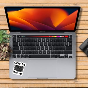 May include: A laptop computer with a sticker that says "Lets do shots!" with a camera icon. The laptop is on a wooden surface with a camera and two potted plants.