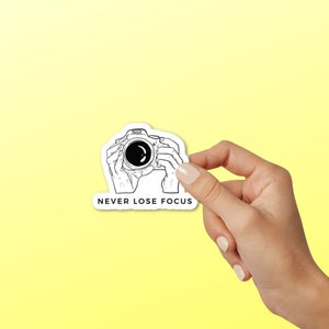 May include: A white sticker with a black line drawing of a camera being held by two hands. The text "NEVER LOSE FOCUS" is below the camera.