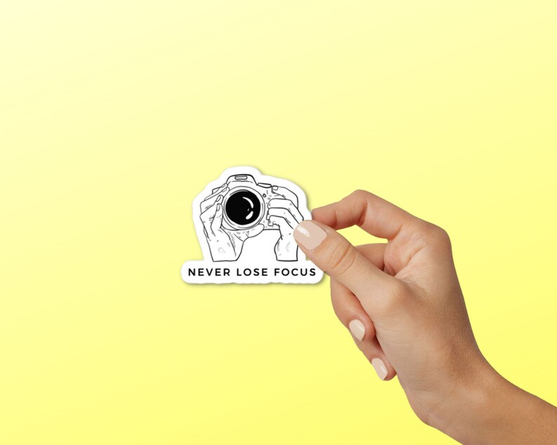 May include: A white sticker with a black line drawing of a camera being held by two hands. The text "NEVER LOSE FOCUS" is below the camera.