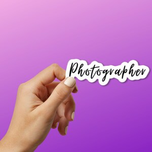 May include: A hand holding a white sticker with the word "Photographer" written in black cursive.