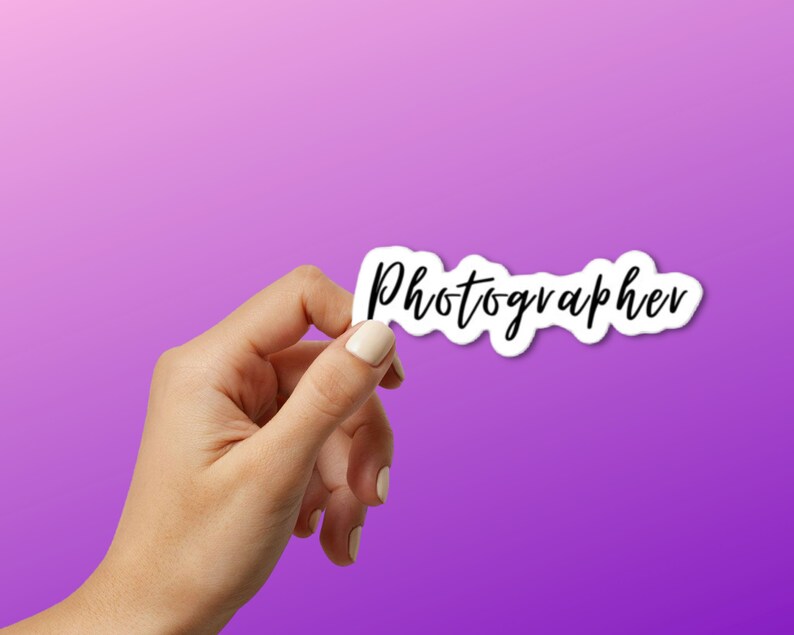 May include: A hand holding a white sticker with the word "Photographer" written in black cursive.
