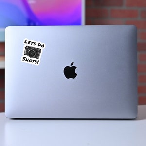 May include: A silver laptop computer with a black Apple logo and a sticker that says "Let's do shots!" with a cartoon camera image.