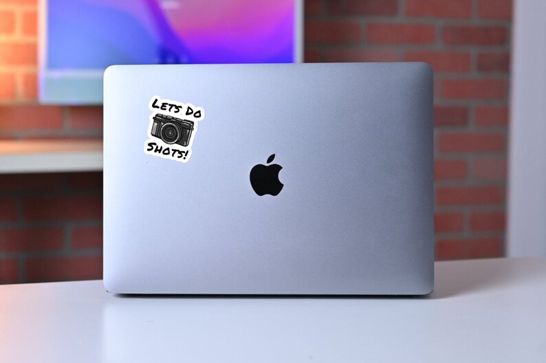 May include: A silver laptop computer with a black Apple logo and a sticker that says "Let's do shots!" with a cartoon camera image.