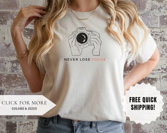 May include: White t-shirt with a black line drawing of a camera and the text "NEVER LOSE FOCUS".