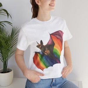 May include: White t-shirt featuring a colorful illustration of a bat with rainbow-colored wings. The bat has a horn and is set against a white background. The shirt is made of a soft material and has short sleeves.
