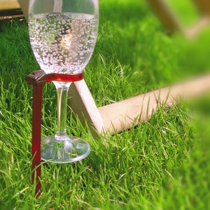May include: A glass of sparkling water is held in place by a red metal holder, which is anchored into the green grass.