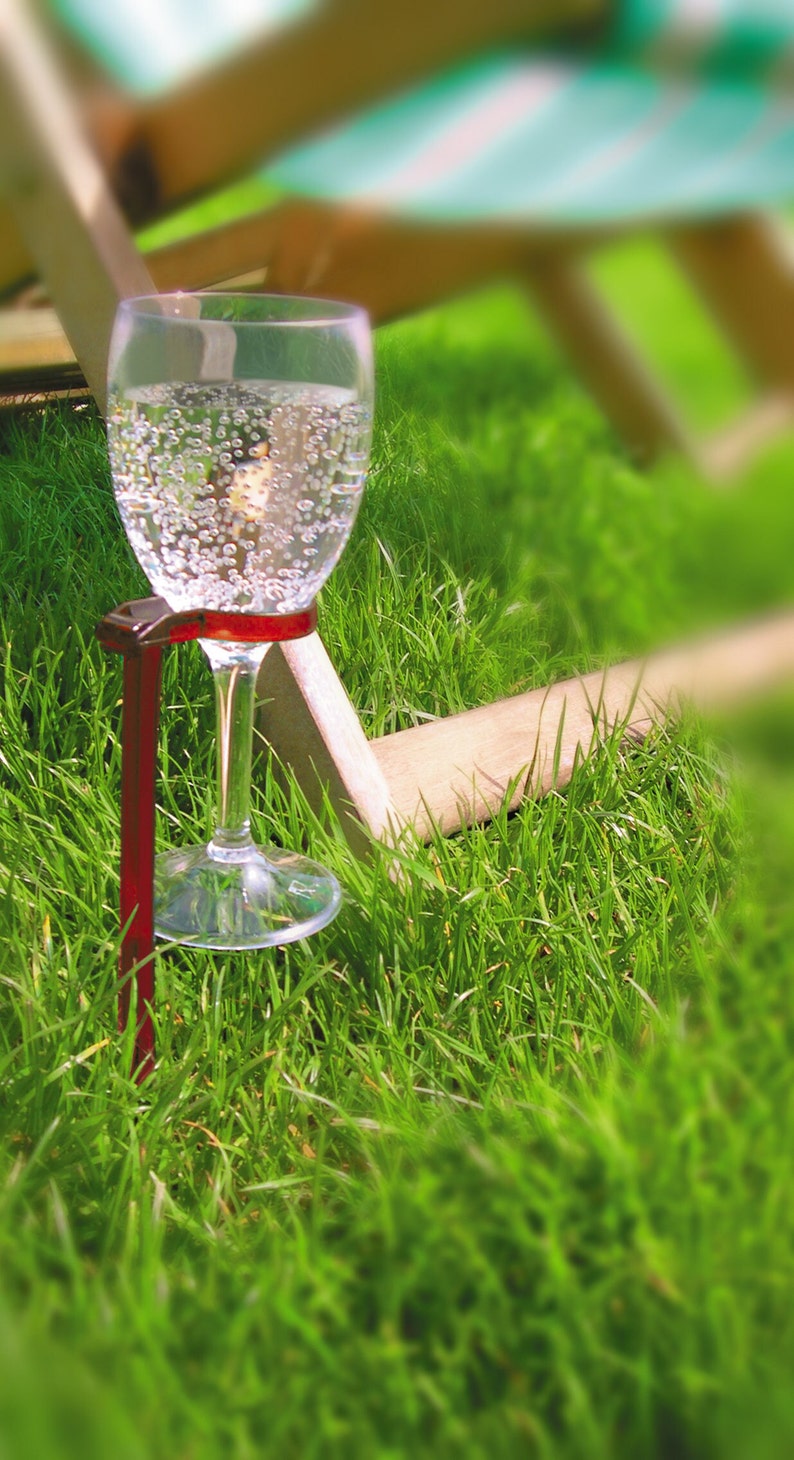 May include: A glass of sparkling water is held in place by a red metal holder, which is anchored into the green grass.