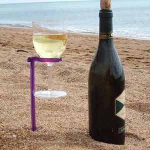 May include: A purple wine glass holder with a glass of white wine and a bottle of wine on a sandy beach.