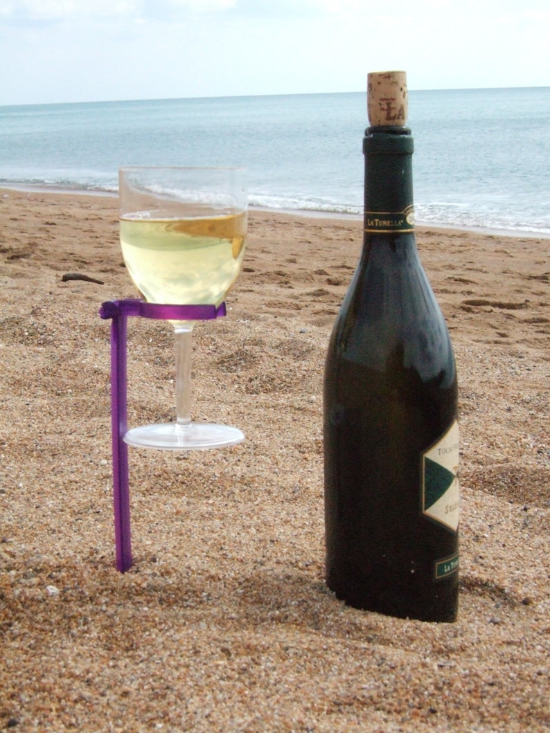 May include: A purple wine glass holder with a glass of white wine and a bottle of wine on a sandy beach.