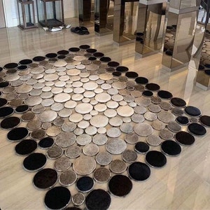Puede incluir: Una alfombra grande, redonda, gris y negra hecha de círculos de cuero. La alfombra está colocada en un suelo claro en una casa moderna.