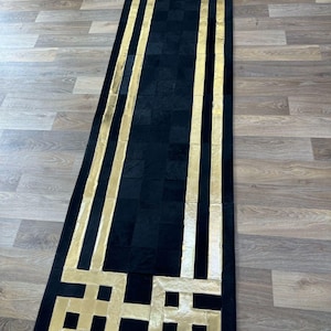 Pode incluir: Um tapete retangular longo com um padrão geométrico em preto e dourado. O tapete é feito de couro e tem um design moderno.