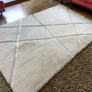 Puede incluir: Una alfombra de pelo blanco con un patrón geométrico de líneas que se cruzan. La alfombra tiene un diseño moderno y está hecha de un material suave y mullido.