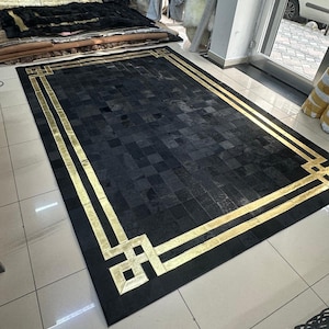 May include: Black and gold patchwork cowhide rug with a geometric border. The rug is made of different pieces of cowhide sewn together to create a unique pattern.
