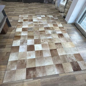 Puede incluir: Una alfombra rectangular grande compuesta por numerosos parches cuadrados en tonos marrones, beige y crema. La alfombra tiene un diseño de patchwork, creando una apariencia texturizada. La alfombra está colocada sobre un suelo de madera.