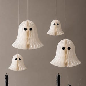 Op de afbeelding: Drie hangende gebroken witte papieren honingraat spookdecoraties met zwarte ronde ogen, opgehangen aan dunne witte koorden, boven zes zwarte taps toelopende kaarsen in zilveren metalen kandelaars, sommige met smeltend kaarsvet en rook.